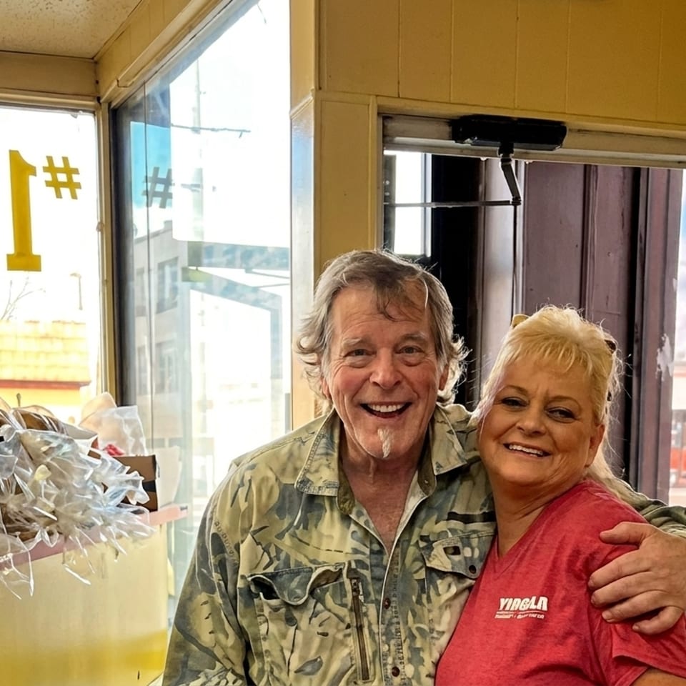 A smiling man and woman embrace inside a bakery, with bread loaves in plastic bags visible.