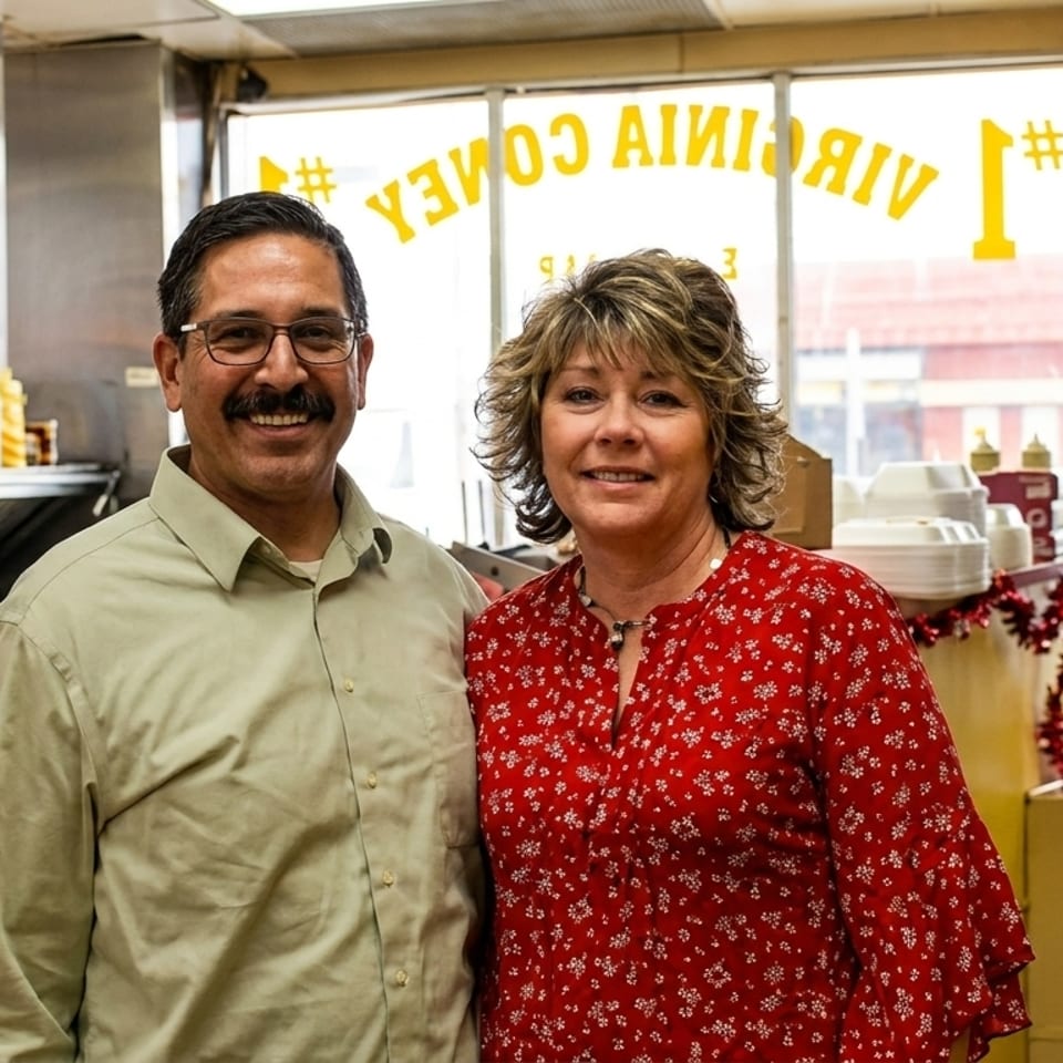 Smiling man and woman standing in a restaurant kitchen with a large window behind them.